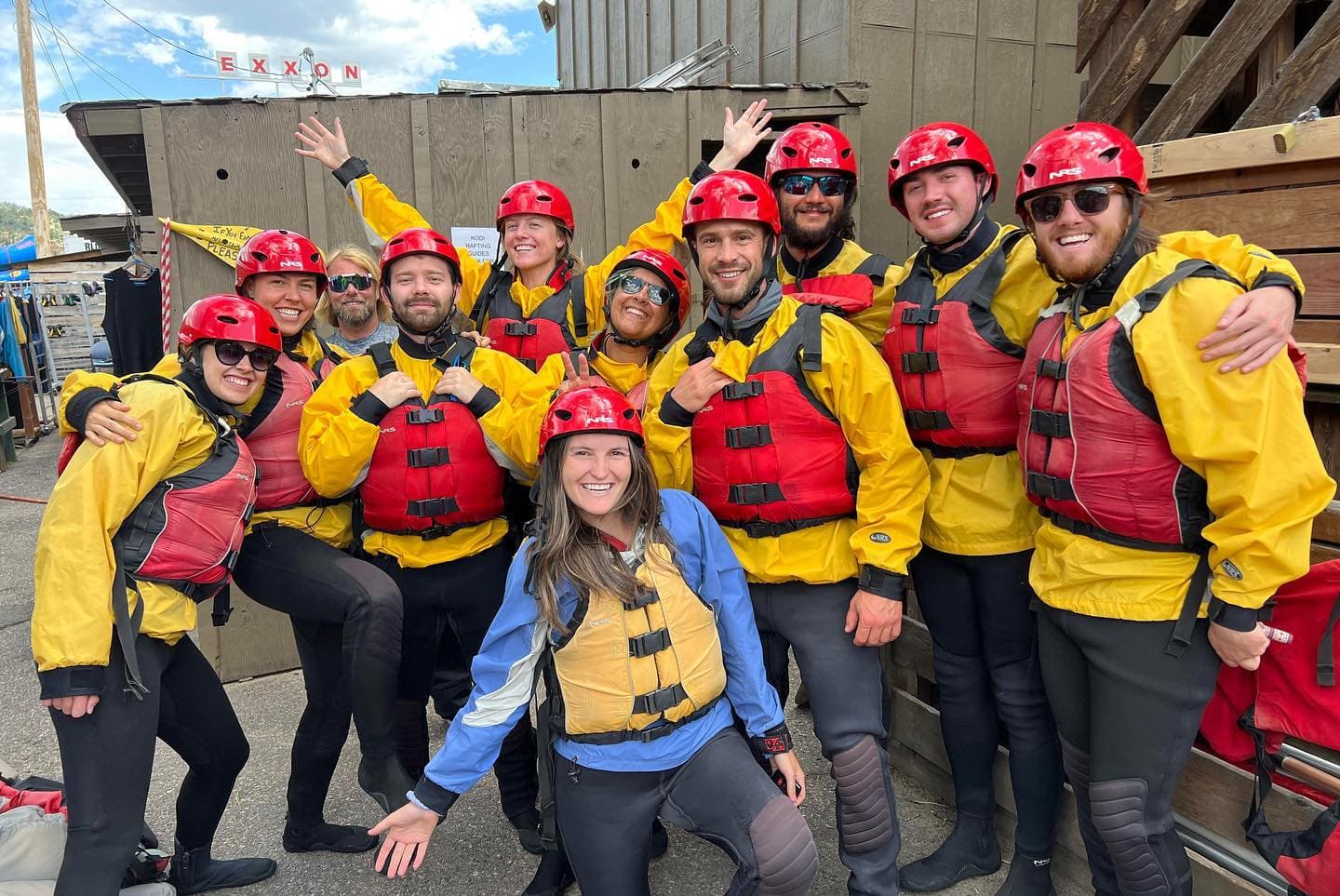 Group of eleven people in yellow rain jackets, red helmets, and life vests posing before rafting